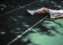 Persona con vestido blanco y zapatillas blancas tumbada en una pista de tenis con luz moteada