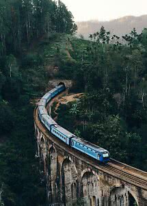 Blue train on a curved stone viaduct through lush forest toward a tunnel in the hills