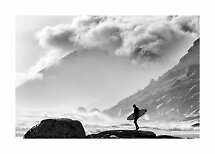 Black-and-white image of surfer with board on rocky shore, waves and misty mountains
