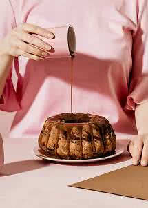 Person in pink blouse pouring chocolate glaze over a marble bundt cake on a plate