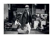 Black-and-white photo of stylish man in coat, cap and sunglasses carrying a large leather bag