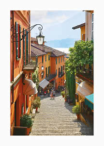 Narrow cobblestone street with colorful houses, balconies, and a lake and mountain view