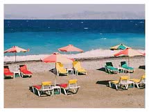 Bunte Sonnenschirme und Liegen am Strand vor türkisblauem Meer mit Bergen im Hintergrund