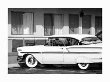 Foto en blanco y negro de un coche clásico blanco aparcado frente a un motel con cortinas.
