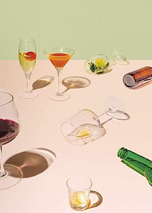 Tisch mit Cocktailgläsern, Wein, Shots und Flaschen auf pastellfarbener Fläche im Sonnenlicht