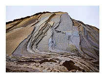 Eine gefaltete Felswand in Sand, Lilagrau und Rost, die Bänder diagonal, oben ein schmales Grasband