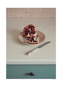 Photograph of an open pomegranate on a floral vintage saucer with a silver knife, on a pale kitchen counter