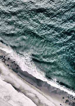 Aerial view of waves crashing onto a dark sandy beach with black rocks