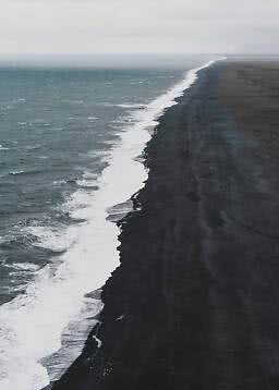 Luftfoto af bølger langs en lang sort sandstrand under overskyet himmel