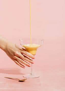 Main tenant un verre à cocktail pendant qu’un dessert orange crémeux y est versé sur fond rose