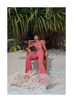 Woman in patterned bandeau top and pants sits on a chair on the beach with waves at her feet