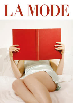 Person in a light blue slip dress lying on a bed, holding a large red book over the face