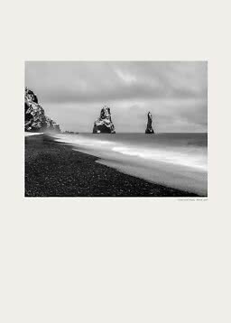 Black-and-white photo of a black sand beach with sea stacks and waves under a cloudy sky