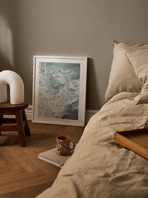 Hyggeligt soveværelse med beige sengetøj, træskammel, hvid lampe og indrammet kunst på gulvet