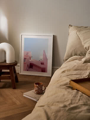 Mysigt sovrum med beige sängkläder, vit bordslampa på pall och inramad rosa konstbild