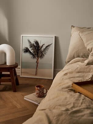 Schlafzimmer mit beiger Bettwäsche, Holzhocker mit weißer Lampe und gerahmtem Palmenbild