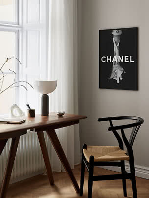 Stilren arbetsplats med träskrivbord, svart stol, bordslampa och inramad Chanel-affisch på väggen