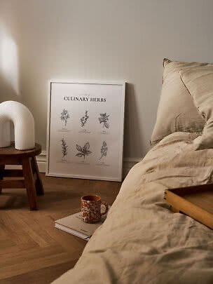Poster incorniciato di erbe aromatiche accanto al letto, con lampada su sgabello e tazza a terra