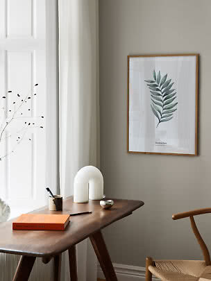 Framed botanical leaf print above a wooden desk in a bright, minimalist room corner