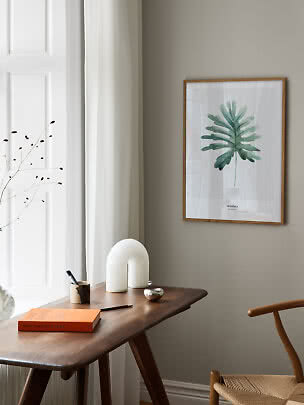 Indrammet plakat med grøn monstera-blad over et træskrivebord i et lyst, minimalistisk rum