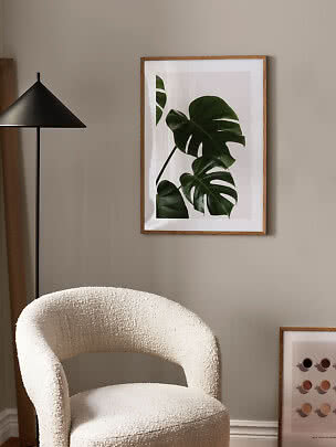 Framed botanical print with green monstera leaves on a beige wall above a cream textured chair.