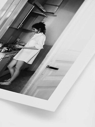 Black-and-white photo of a barefoot woman in an oversized shirt cooking in a small kitchen