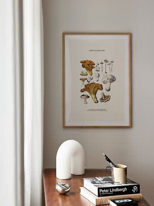 Framed botanical mushroom print above a wooden table with books, mug, and white sculptural lamp