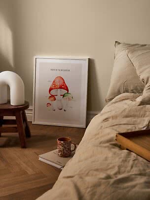 Framed mushroom poster leaning by bed, with white lamp on stool and mug on a book