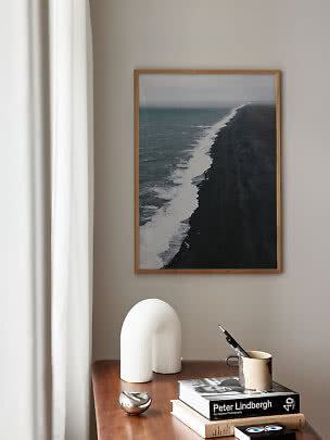Indrammet kystfotografi over sort sandstrand på beige væg over et træbord med bøger og dekoration