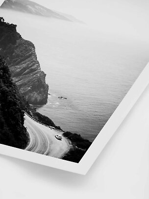 Black-and-white photo of a coastal road by cliffs with a car beside the sea