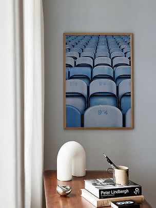 Indrammet plakat med rækker af blå stadionsæder på grå væg over et træbord