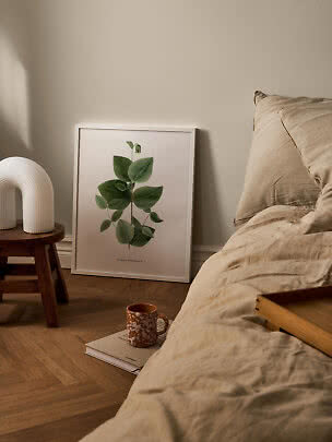 Indrammet botanisk plakat lænet op ad væg ved seng med beige sengetøj og træbord