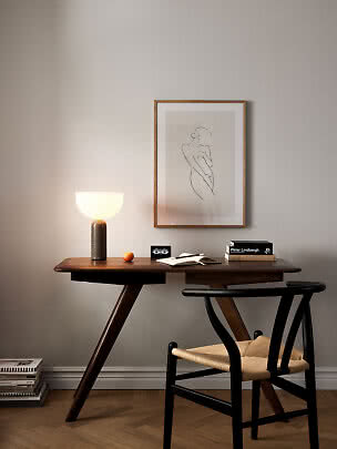 Dark wood desk with glowing table lamp, books, open notebook and black chair beneath framed line art.