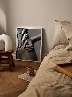 Cozy bedroom with beige bedding, stool with white lamp, and black-and-white framed print on the floor