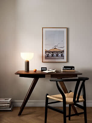 Mørkt træskrivebord med sort stol, tændt bordlampe og indrammet plakat på beige væg