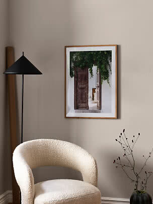 Framed poster of a rustic doorway with greenery, hung on a beige wall above a cream armchair