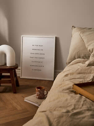 Minimalistisches Schlafzimmer mit beiger Bettwäsche, gerahmtem Spruchposter, weißer Lampe und Tasse auf Buch
