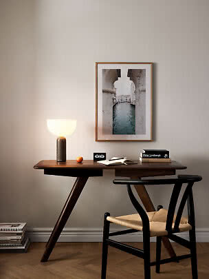 Dark wooden desk with table lamp, books, notebook and black chair beside a framed canal print.