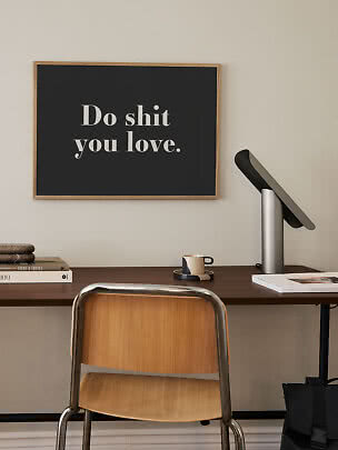 Sort typografiplakat i træramme med teksten "Do shit you love" over et skrivebord.