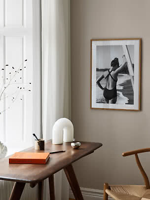 Træindrammet sort-hvid plakat over et skrivebord med bog, vase og hvid bordlampe.