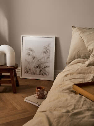 Cozy bedroom with beige bedding, white lamp on a stool, and framed grass print on the floor