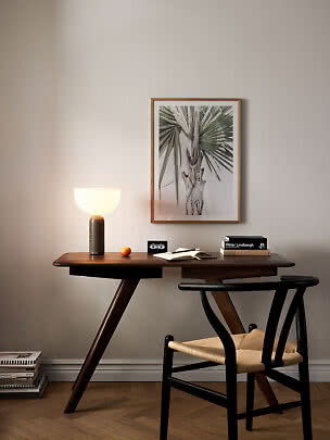 Mørkt træskrivebord med tændt bordlampe, stol og indrammet palmeprint på væggen.