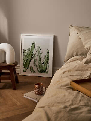 Ingelijste cactusposter leunt tegen de muur naast een bed en houten kruk in een warme slaapkamer