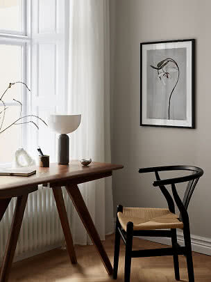 Minimalistisk hjemmekontor med træbord, sort stol, bordlampe og botanisk plakat ved vindue.