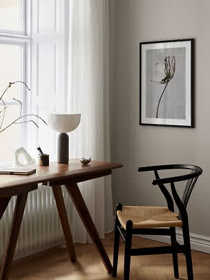 Minimalistischer Arbeitsplatz mit Holztisch, schwarzem Stuhl, Tischlampe und gerahmtem Blumenbild.