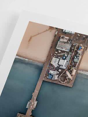 Angled aerial photo print of a pier with rides beside a sandy beach and ocean, with white border