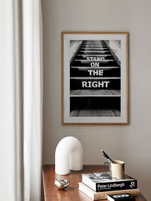 Ingelijste zwart-wit poster met roltrap en tekst "STAND ON THE RIGHT" aan beige muur