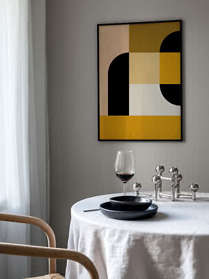 Abstrakt poster med gula, beige, vita och svarta former i svart ram ovanför ett dukat bord.