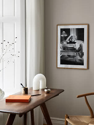 Framed black-and-white photo on beige wall above wooden desk with white lamp and book