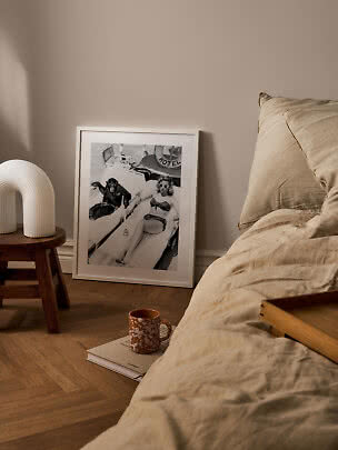 Cozy bedroom with beige bedding, wooden stool, white lamp and framed black-and-white poster